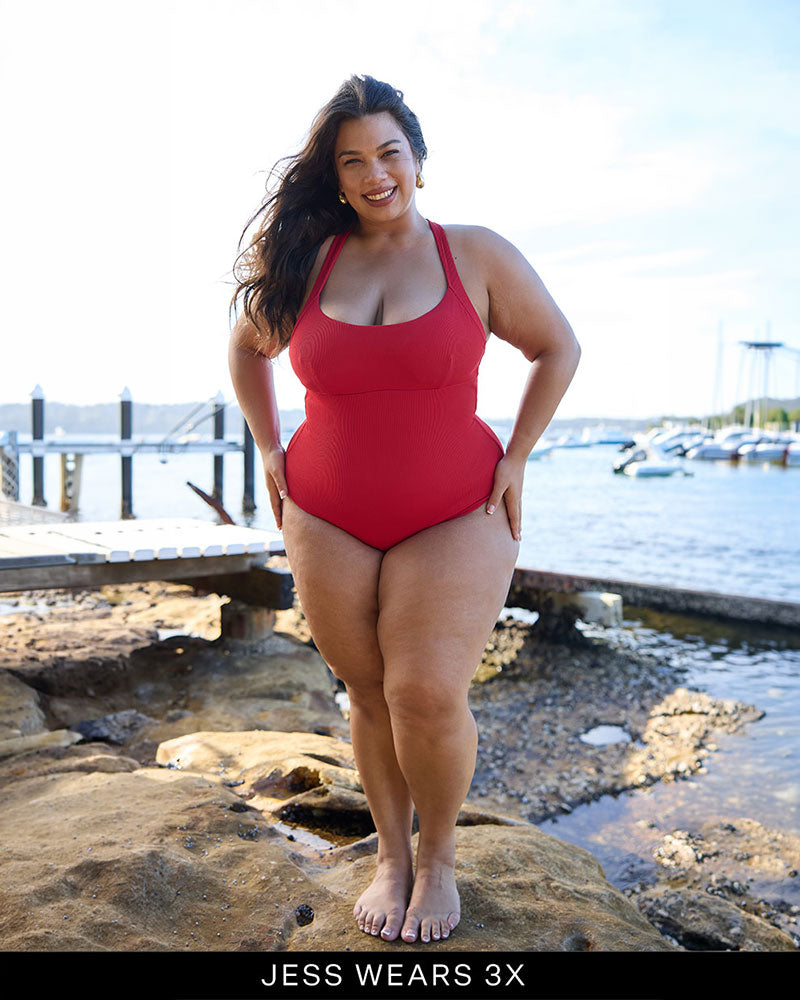 Woman in a Fearless red supportive bust and one piece swimsuit standing by a waterfront, sunlit background