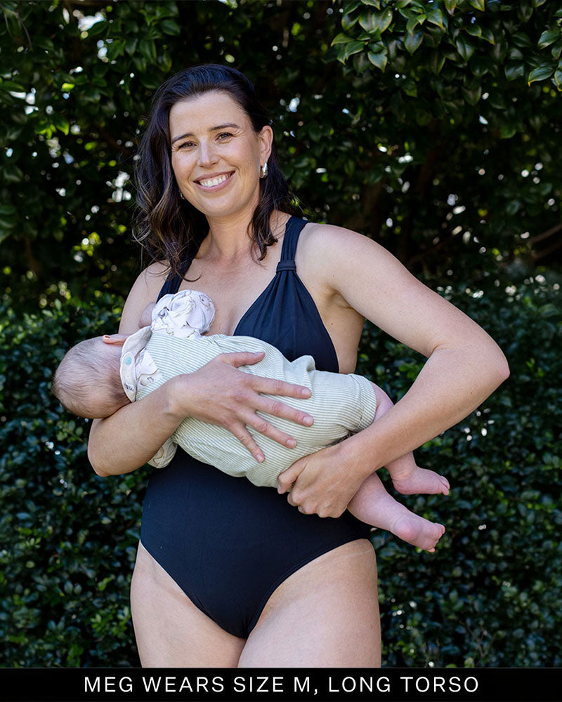 Woman in a black postpartum supportive bust swimsuit holding a baby against a green leafy background.
