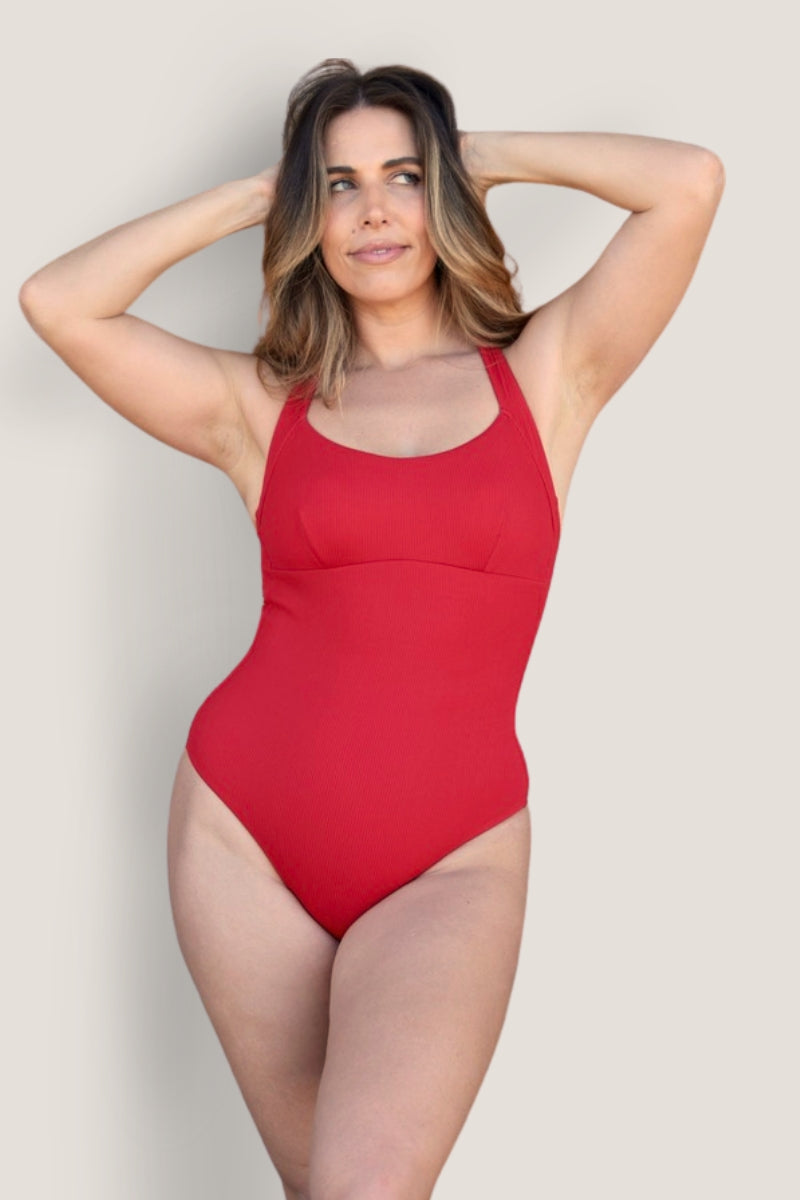A woman with wavy, shoulder-length hair poses in the Fearless au Adventure Suit, a red swimsuit made from recycled ribbed fabric, against a plain light background with her arms raised and hands resting on her head.