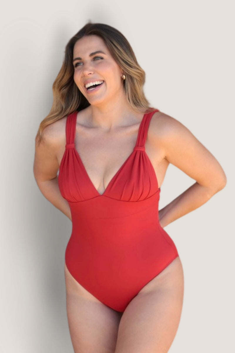A smiling woman with long, wavy hair wears the Fearless au All Day One Piece - Standard, a red swimsuit with tummy-hugging compression and gathered straps, standing against a plain light background.