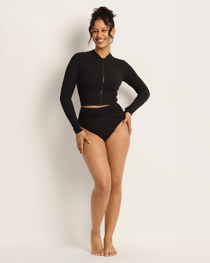 Woman wearing a cropped black UPF rash vest and black full coverage tummy control bikini bottoms standing in front of a white background.