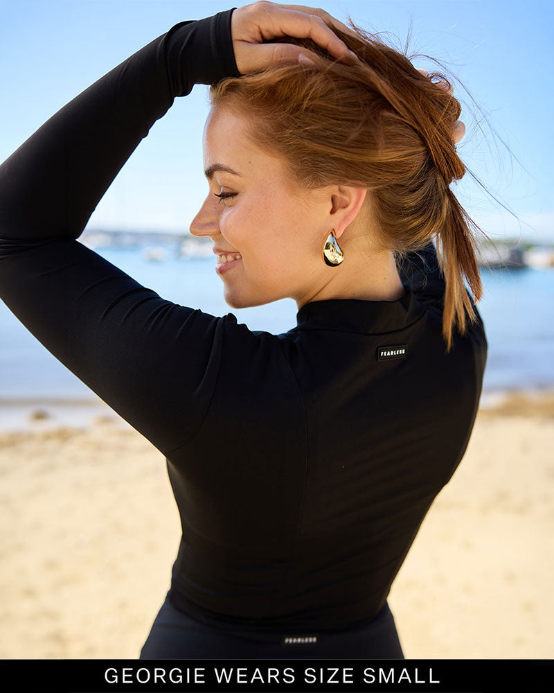 Woman wearing a black UPF rash vest on a beach with water and sky in the background