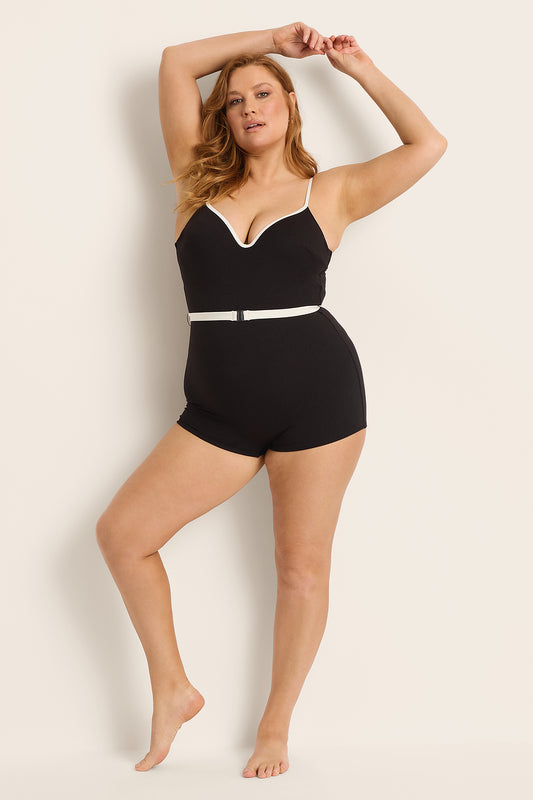 A woman poses barefoot against a beige background wearing the Fearless au Grace Boyleg swimsuit in Matte Black / Standard, featuring white trim and a white belt, with one arm raised above her head.