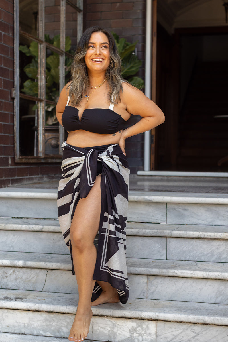 A woman in the Fearless au Geo Print Sarong and a black bikini top smiles as she walks barefoot down marble steps, surrounded by brick walls and lush green plants.