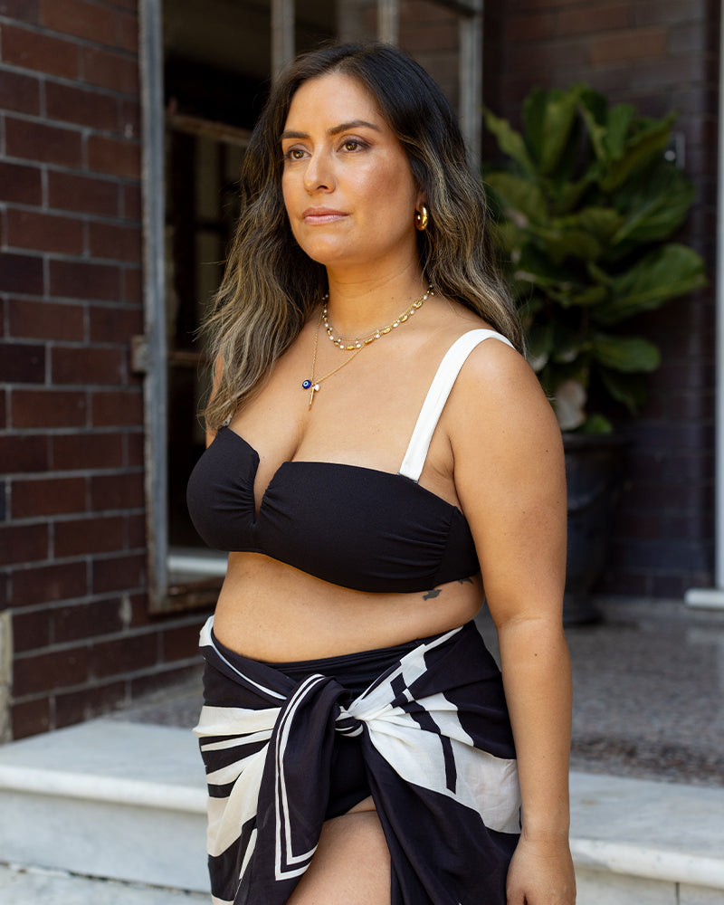A woman with long brown hair stands outside wearing a black bikini top with white straps and a black-and-white sarong. She has gold jewelry and looks ahead with a neutral expression, with brick walls and plants in the background.