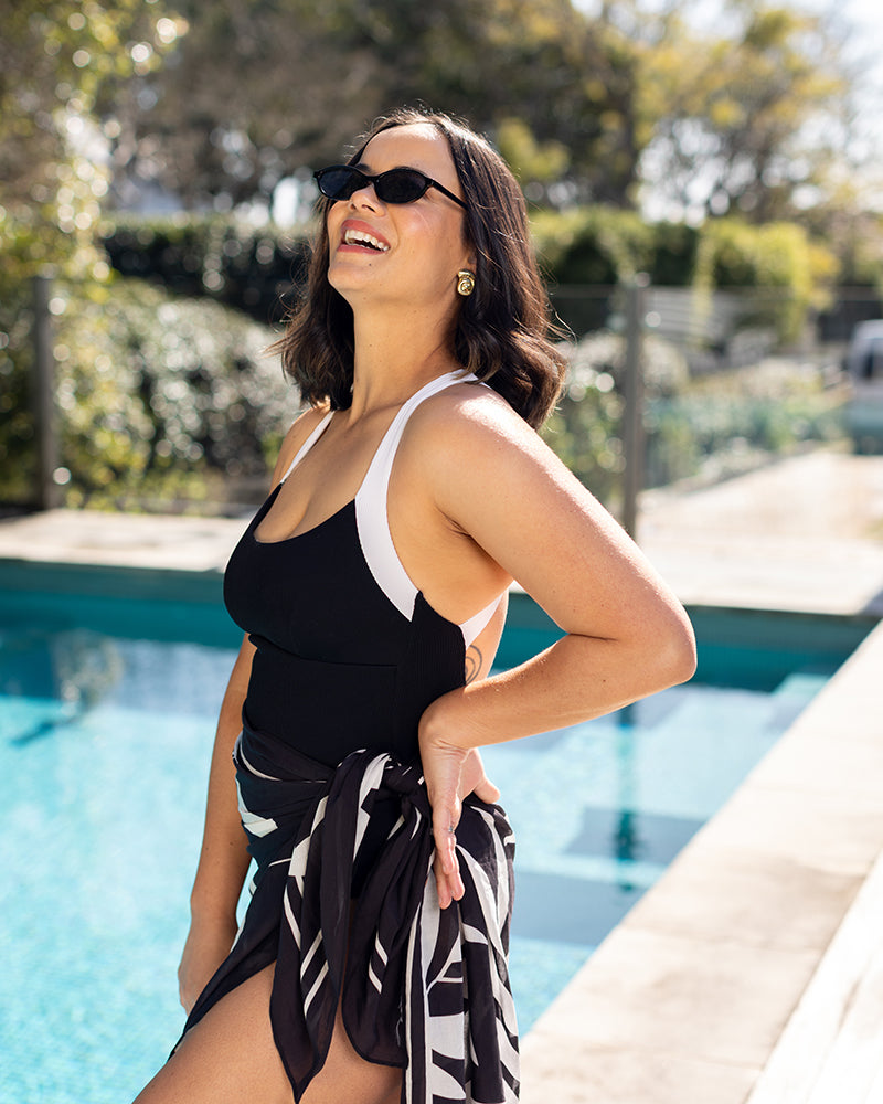 A woman wearing sunglasses, a black and white swimsuit, and a patterned sarong stands by a swimming pool in the sunlight, smiling and looking up. Trees and greenery are visible in the background.