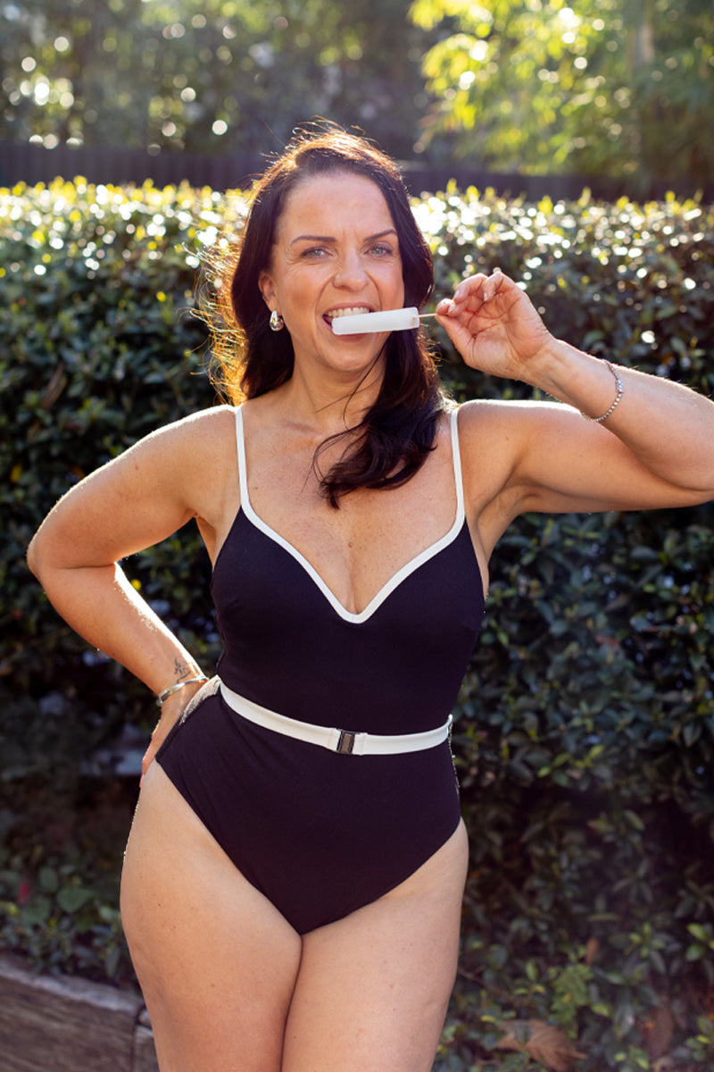 A woman confidently poses outdoors in the Fearless au Grace High Rise - Matte Black one-piece, smiling with a tampon between her teeth, surrounded by sunlit trees and green shrubbery.