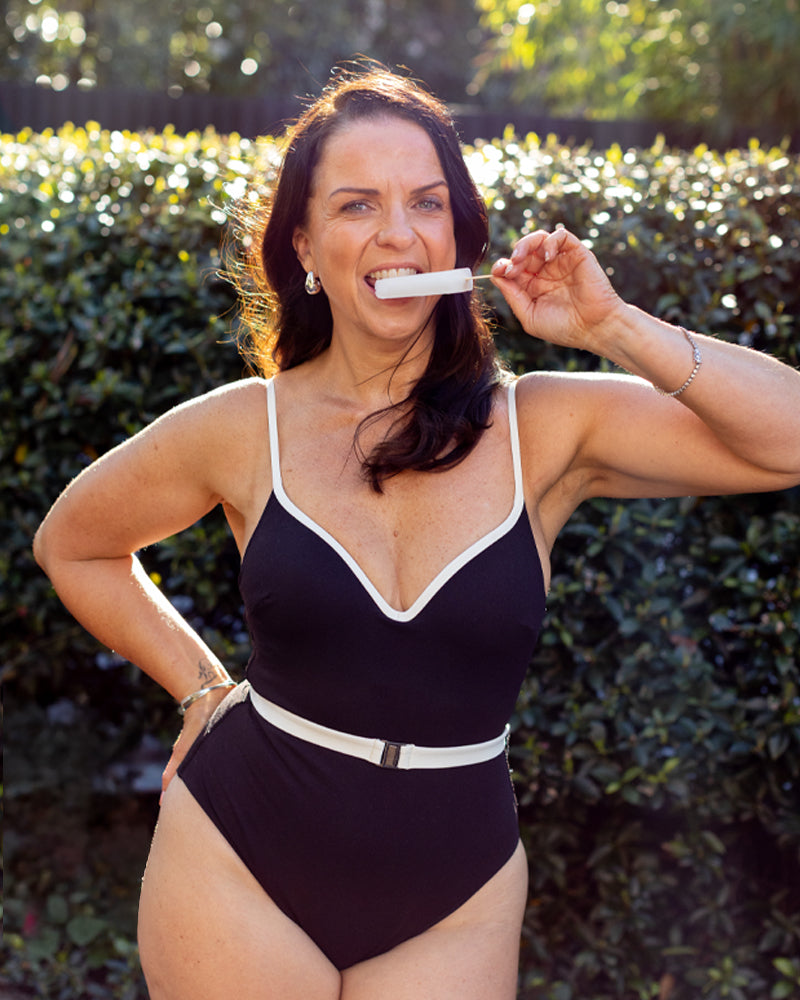 A woman in a black one-piece swimsuit stands outdoors in front of green bushes, smiling and holding a popsicle to her mouth in the sunlight.