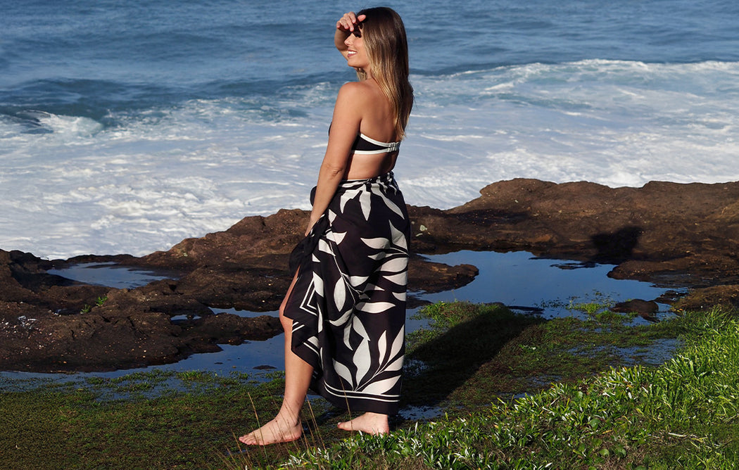 A woman in a black and white patterned wrap skirt and bikini top stands barefoot on a rocky, grassy shore, smiling and looking towards the ocean waves in the background.