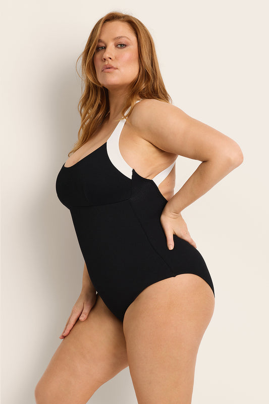 A woman with long, reddish-brown hair poses confidently in the Fearless au Adventure Suit Long Torso, a recycled ribbed swimsuit with white shoulder straps and a built-in shelf bra, against a plain light background.