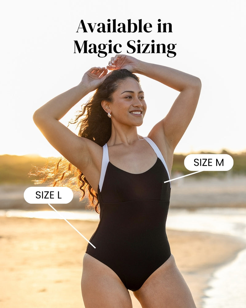 A smiling woman stands on a beach wearing a black one-piece swimsuit with white straps. Text reads Available in Magic Sizing with arrows pointing to the swimsuit marked Size L and Size M.
