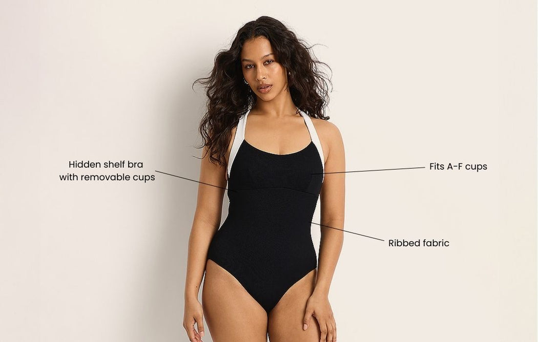 A woman wears a black one-piece swimsuit with white straps. Text points to features: “Hidden shelf bra with removable cups,” “Fits A–F cups,” and “Ribbed fabric.” The background is plain and light-colored.