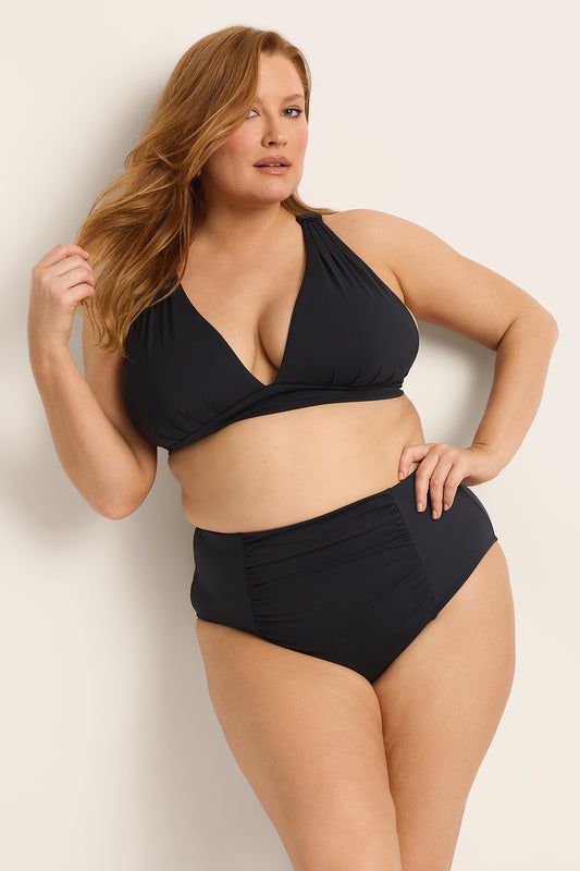 A woman with long, light brown hair confidently models the Fearless au All Day Bikini Bottom in Matte Black, pairing high-waisted tummy control swimwear with a v-neck top against a neutral background.