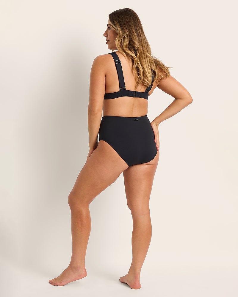 A woman stands barefoot facing away, wearing a black high-waisted bikini bottom and a matching black bikini top with wide straps, posing against a plain light background.