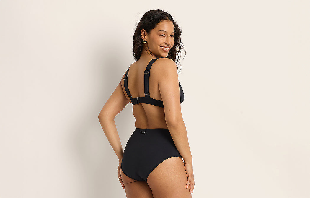 A woman stands smiling and looking over her shoulder, wearing a black high-waisted bikini set. The background is plain and light-colored.