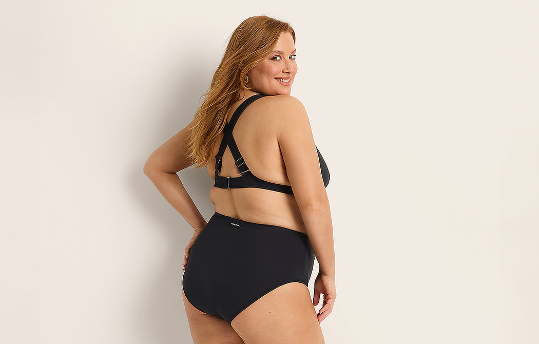 A smiling woman with long red hair models a black high-waisted bikini set, standing sideways with one hand on her hip against a plain light background.