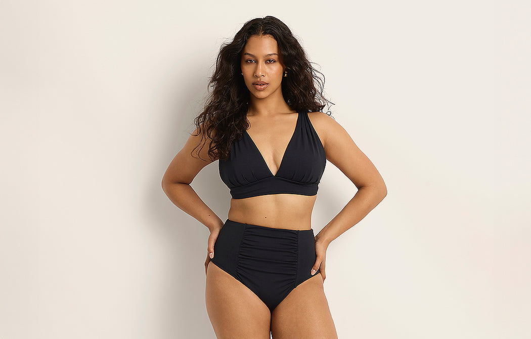 A woman with long, wavy hair wears a black high-waisted bikini with a V-neck top. She stands against a plain, light-colored background with one hand on her hip, looking confidently at the camera.