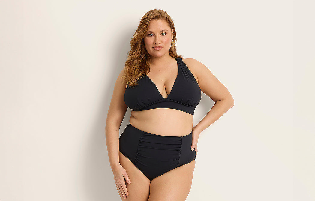 A woman with long, light brown hair poses confidently in a black two-piece bikini with a deep V-neck top and high-waisted bottoms against a plain, light background.