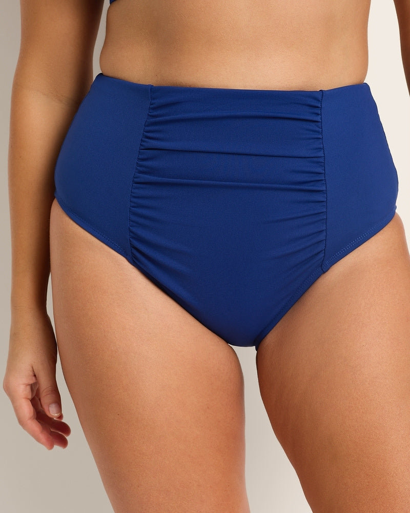 A close-up of a person wearing high-waisted, royal blue bikini bottoms with ruched detailing at the front. The background is neutral and the swimwear fits snugly at the waist and hips.