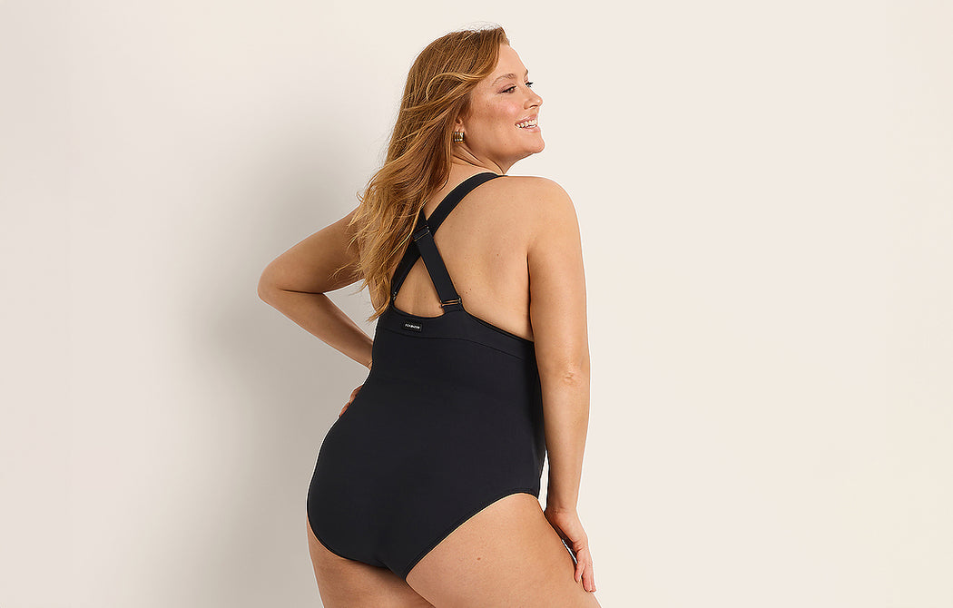 A woman with long hair smiles while posing in a black one-piece swimsuit with a racerback design, standing against a plain light-colored background.