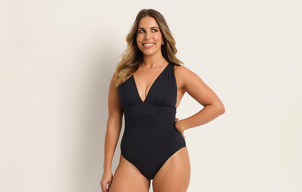 A woman with long, wavy hair is smiling and posing in a black one-piece swimsuit against a plain, light-colored background.