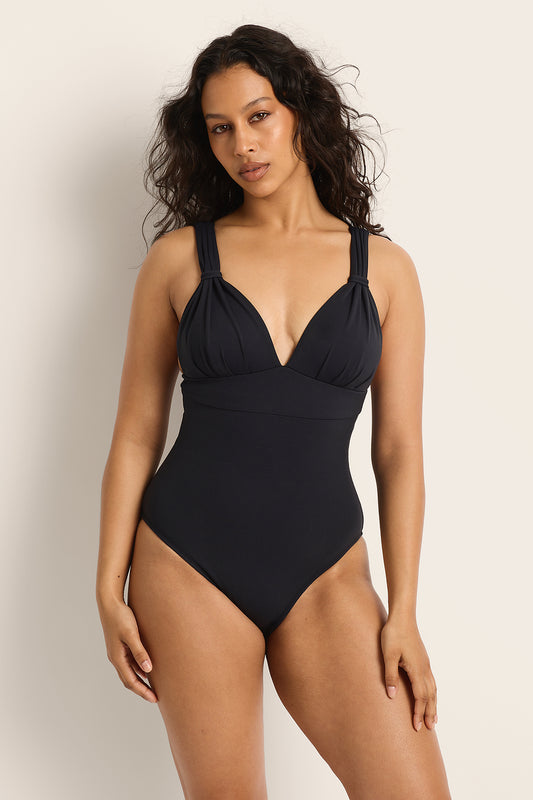 A woman with wavy dark hair models the Fearless au All Day One Piece in Matte Black/Long, featuring thick shoulder straps, built-in bust support, and a deep V-neckline, against a plain light background.
