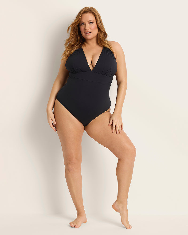 A woman with long, reddish-blonde hair poses confidently in a black one-piece swimsuit against a plain, light background. She is standing barefoot with one leg slightly bent and one hand resting on her hip.