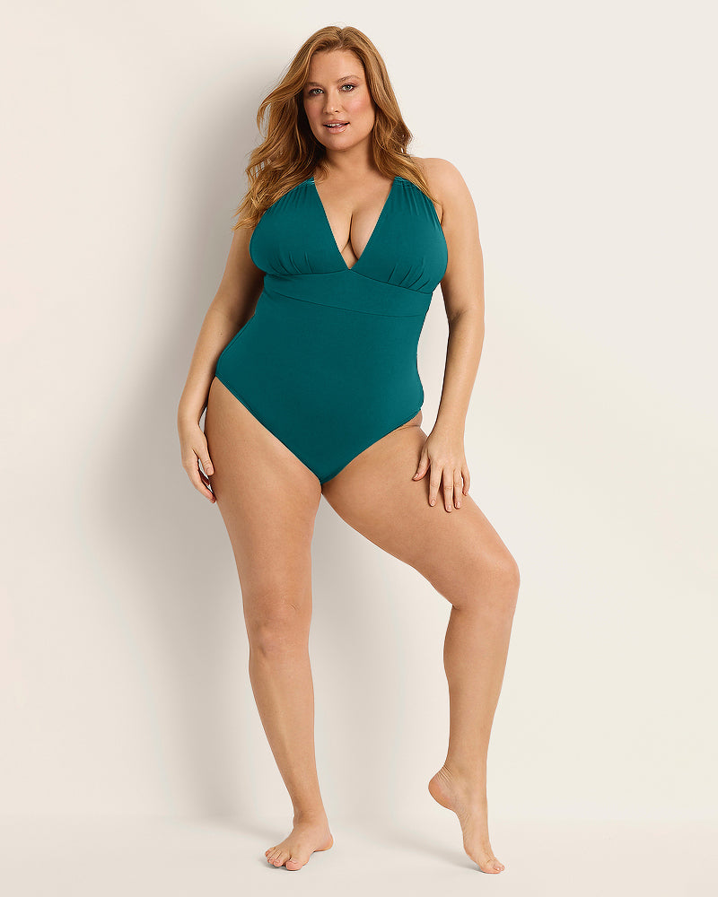 A woman with long red hair poses barefoot with one hand on her hip, wearing the Fearless au All Day One Piece - Standard, a teal tummy control swimsuit featuring a deep V-neckline, against a plain light background.