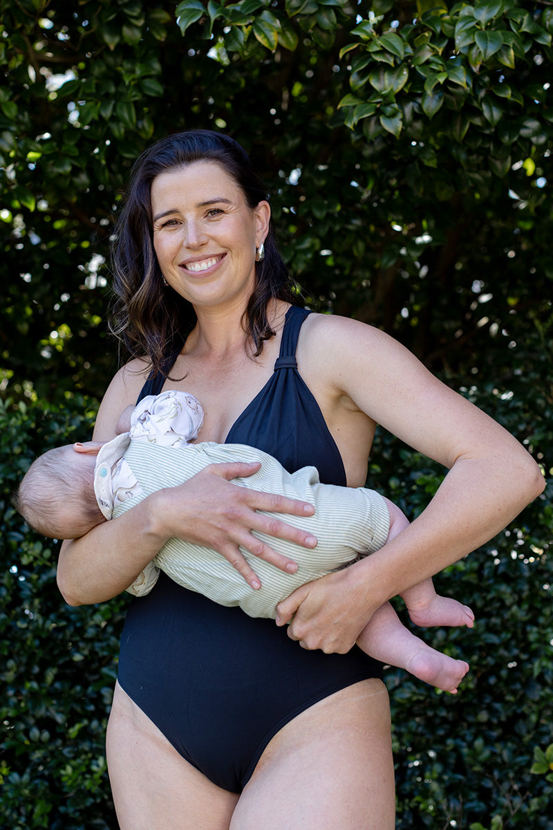 A smiling woman wearing the Fearless au All Day One Piece - Matte Black/Long with built-in bust support stands outdoors with green foliage behind her, holding a baby in her arms.