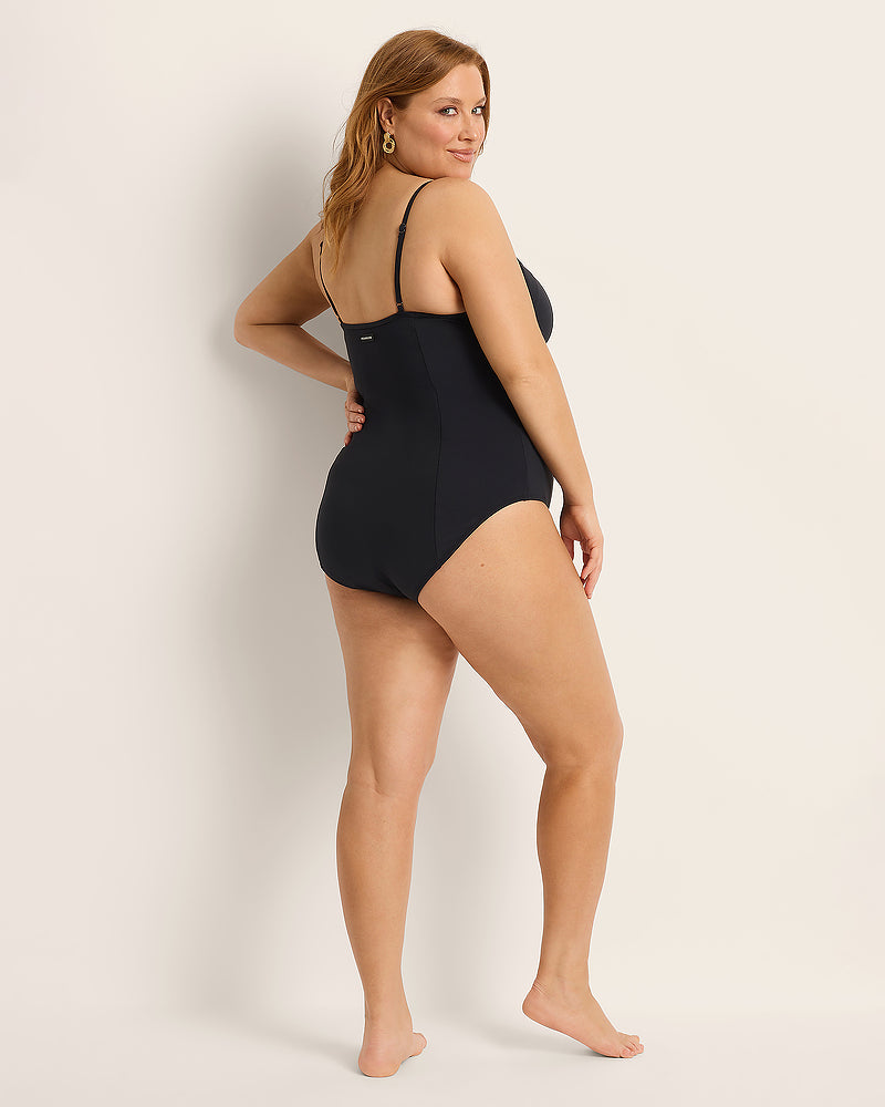 A woman with long hair poses barefoot in a black one-piece swimsuit, standing turned slightly to the side and looking over her shoulder against a plain light background.