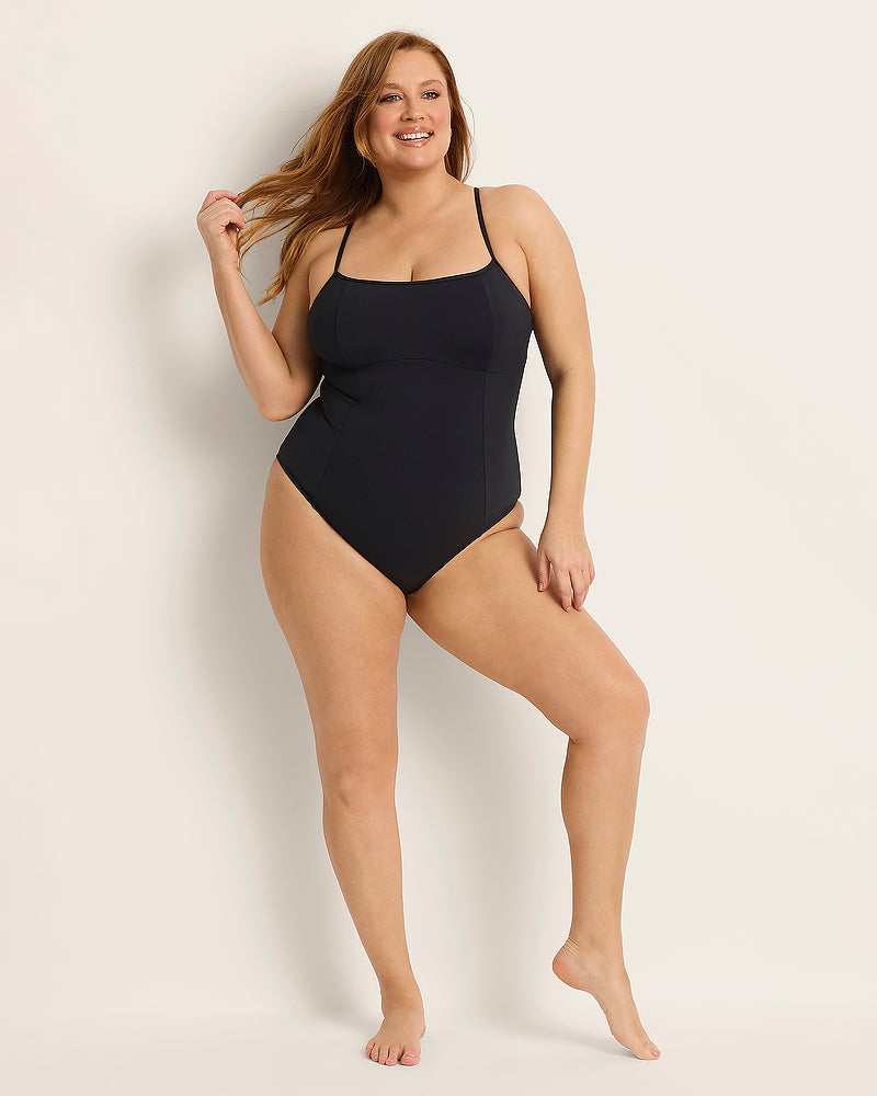 A smiling woman with long, wavy hair poses in a simple, black one-piece swimsuit against a plain, light-colored background. She stands barefoot, holding a strand of her hair, exuding confidence and positivity.
