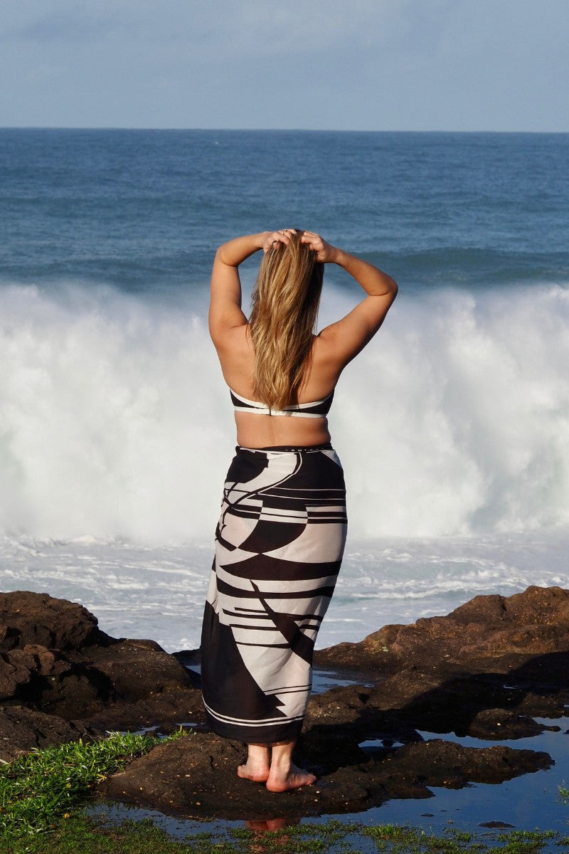 A woman with long hair stands barefoot by the ocean, waves crashing behind her, wearing a Fearless au Geo Print Sarong as a cover-up over her bikini. Her arms are raised, showcasing the vibrant geometric pattern.