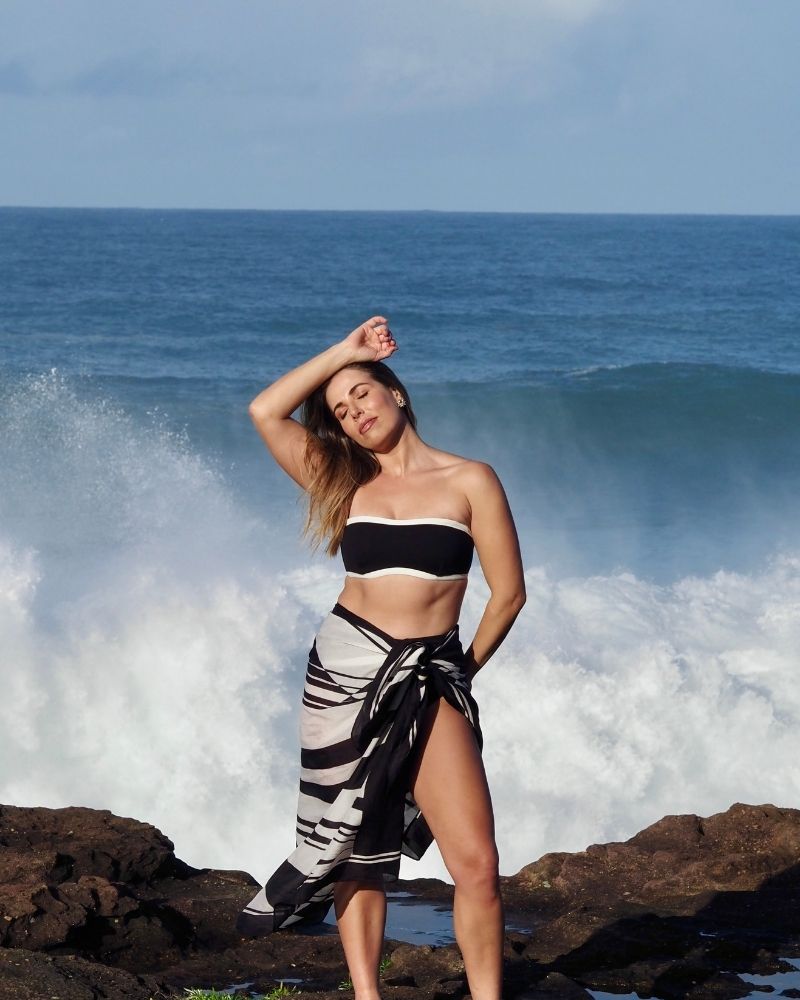 A woman in a black bandeau bikini top and a black-and-white sarong stands on rocks by the ocean, posing with one arm raised and eyes closed, with waves crashing behind her under a blue sky.