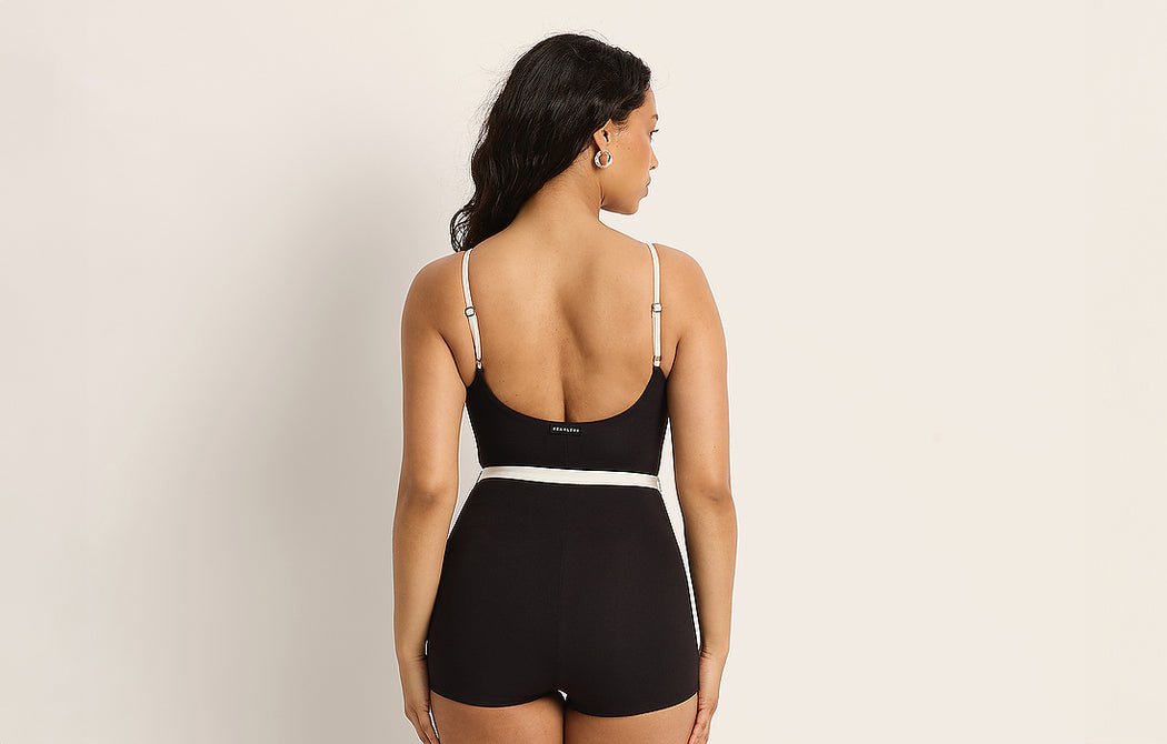 A woman with long dark hair stands facing away, wearing a black sleeveless romper with adjustable straps and a white waistband, against a plain light background.