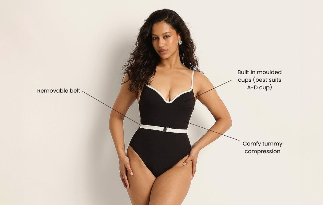 Woman wearing a black one-piece swimsuit with white trim and a removable belt, featuring labels for built-in moulded cups (best for A–D cup) and comfy tummy compression.