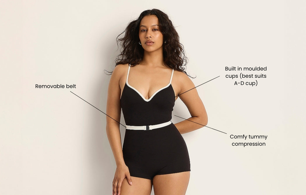 A woman wears a black one-piece swimsuit with white trim and a removable belt. Text points to features: built-in moulded cups (A-D cup) and comfy tummy compression. The background is plain and light-colored.