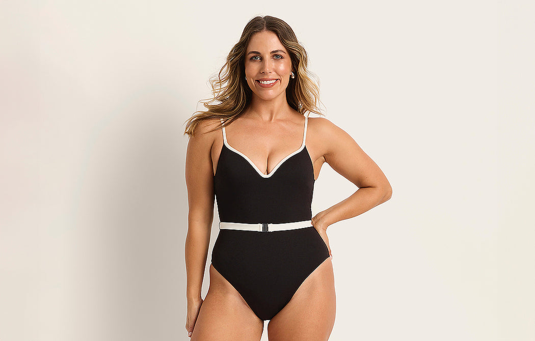A woman with long, wavy hair smiles while wearing a black one-piece swimsuit with white trim and a white belt, posing against a plain light background.