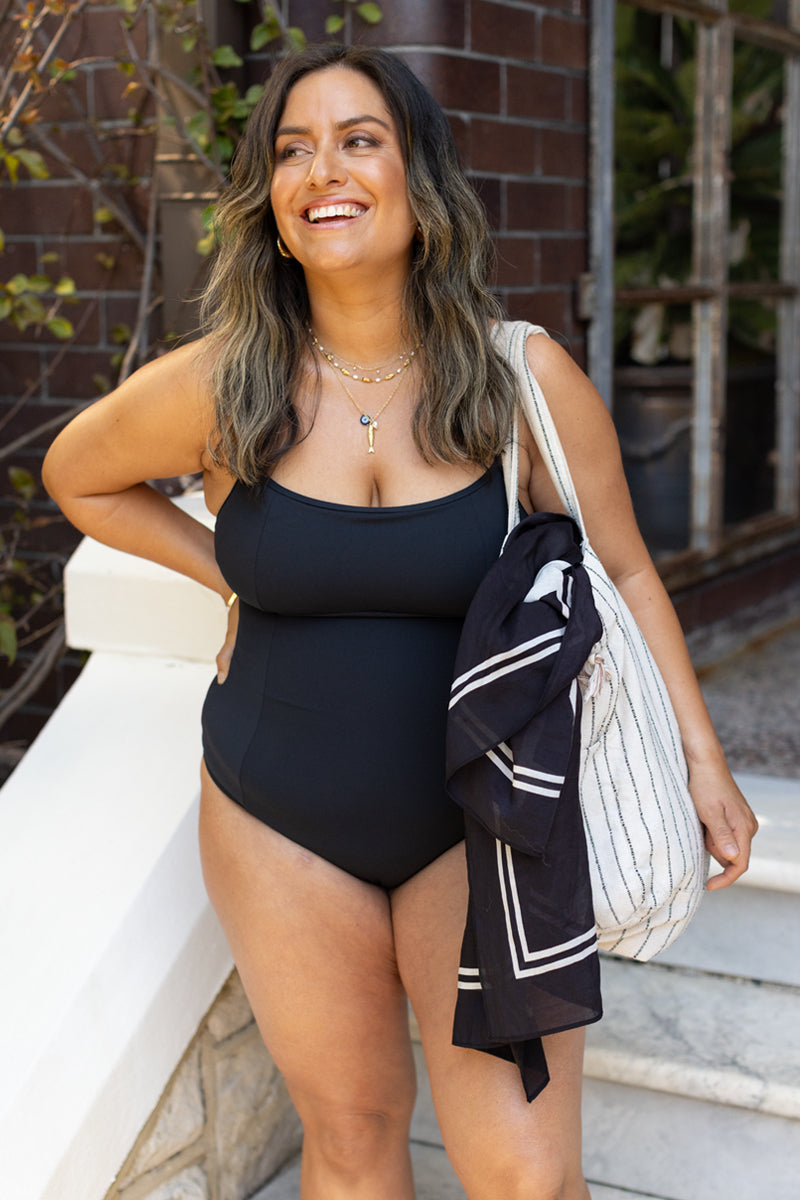 A woman with long dark hair smiles outdoors, wearing the Fearless au Freedom One Piece swimsuit made from recycled fabric, layered necklaces, and holding a striped bag with a scarf tied to it. She looks relaxed and happy.