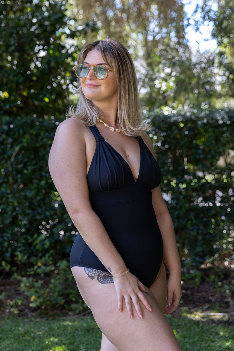 A woman stands outdoors on grass, smiling, wearing Fearless au's All Day One Piece - Matte Black/Long swimsuit with built-in bust support. Trees and greenery are in the background, and she accessorizes with sunglasses.
