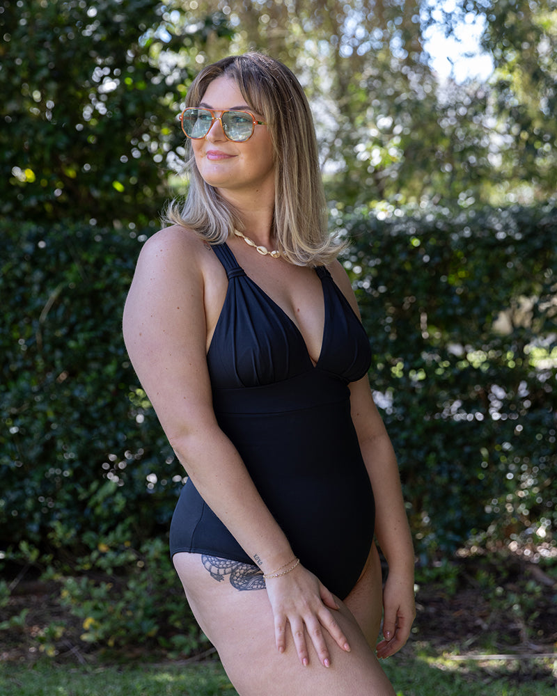 A woman with shoulder-length blonde hair wearing sunglasses and a black one-piece swimsuit stands outdoors in front of green foliage, smiling and looking to the side.