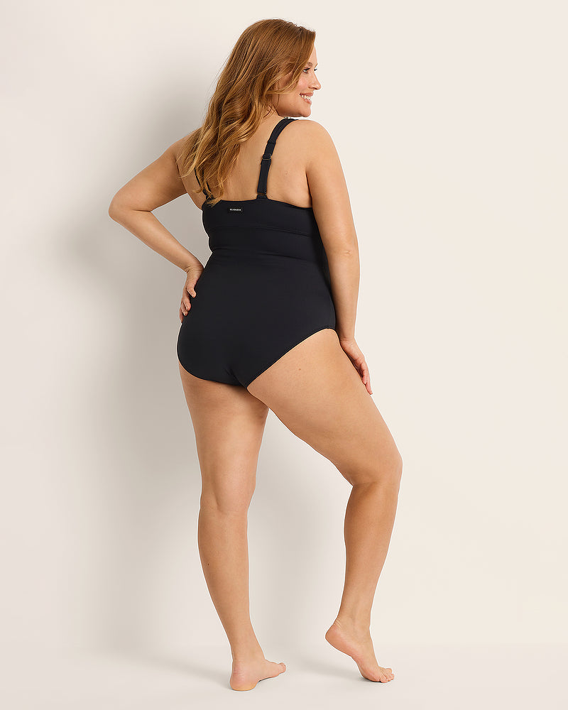 A woman with long, wavy hair stands barefoot on a light background, wearing a black one-piece swimsuit. She is turned slightly to the side, smiling, with one hand resting on her hip.