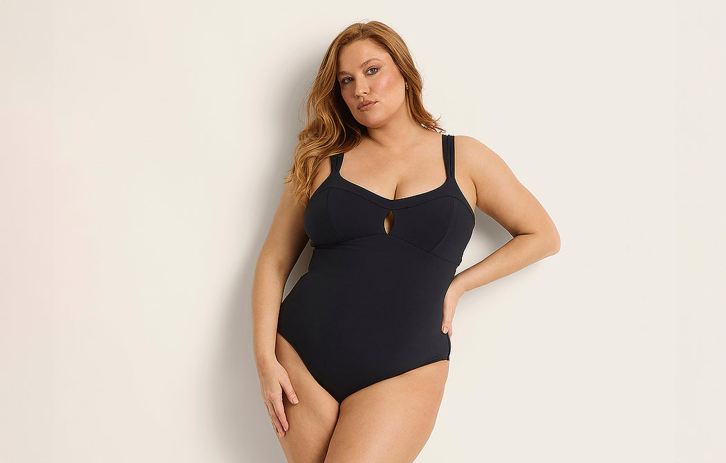 A woman with long, light brown hair wears a black one-piece swimsuit with thick straps and a small front cutout, standing against a plain light-colored background.