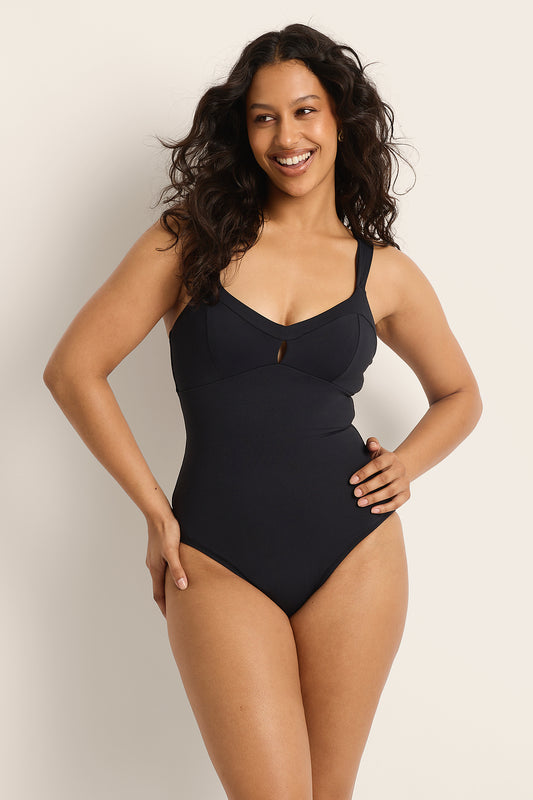 A woman with long, wavy dark hair smiles in the Fearless au Magic Suit - Matte Black / Standard, a supportive swimsuit made from recycled fabric. She poses with one hand on her hip and the other on her thigh against a light background.