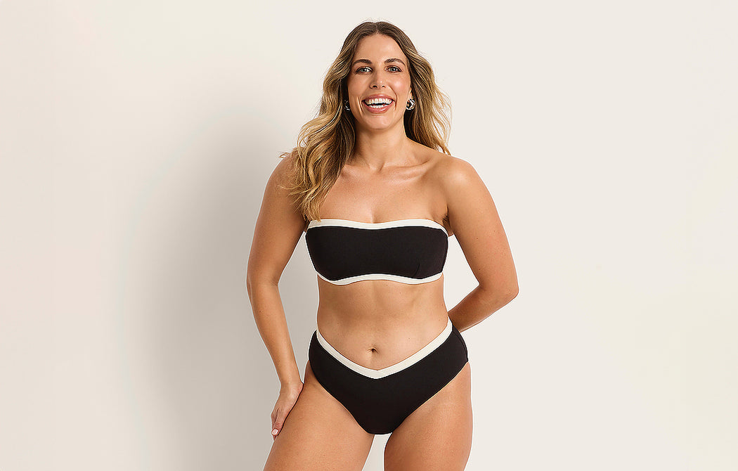 A woman with long, wavy hair smiles while wearing a black strapless bikini with white trim, standing against a plain light background.