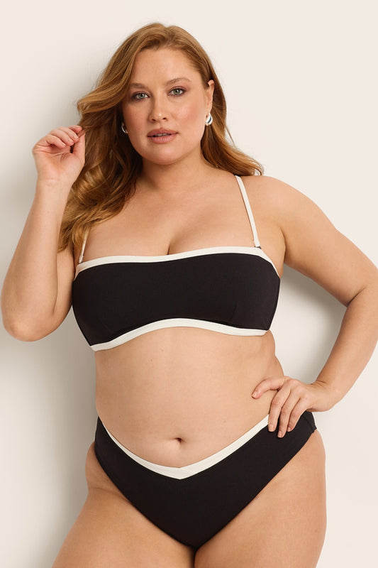 A woman with long, wavy auburn hair confidently models the Fearless au Nova Bikini Top in matte black with white trim, featuring a convertible design, against a neutral background.