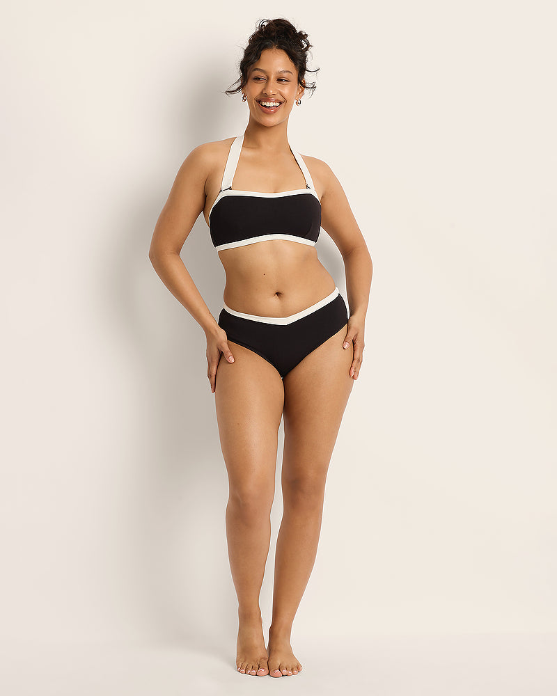 A woman smiles while wearing a black and white halter bikini set, standing barefoot against a plain light background.