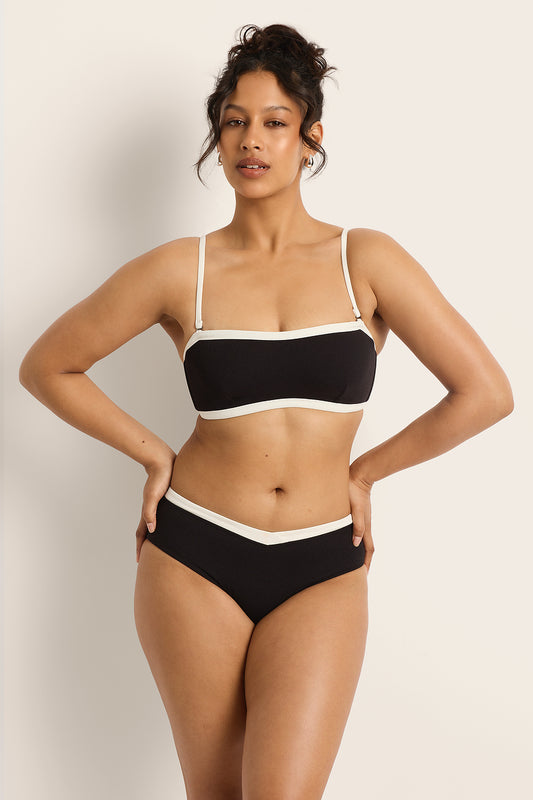 A woman poses confidently with one hand on her hip, wearing the Fearless au Nova Bikini Bottom in matte black with white trim, against a plain beige background.
