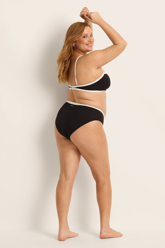 A woman with light skin and long, reddish-blonde hair smiles barefoot against a cream background, posing with arms raised in a V-cut bikini and Fearless au's Nova Bikini Bottom.