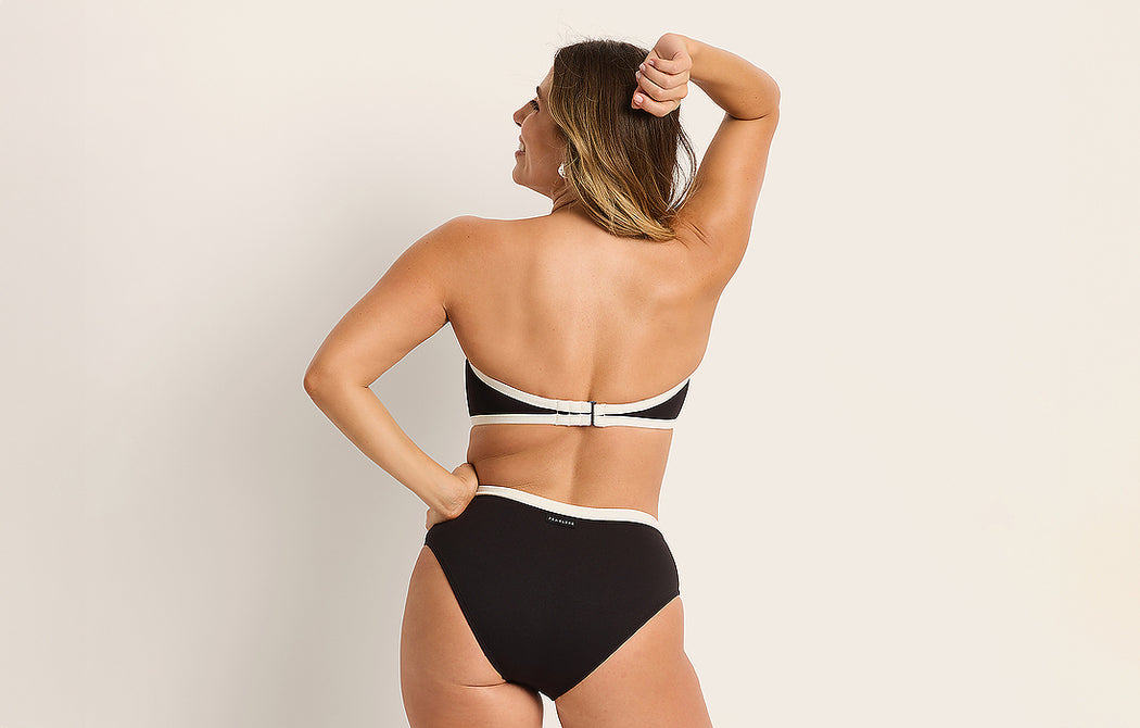 A woman stands with her back to the camera, wearing a black and white strapless bikini. One arm is bent with her hand on her head, and the other rests on her hip. The background is plain and light-colored.