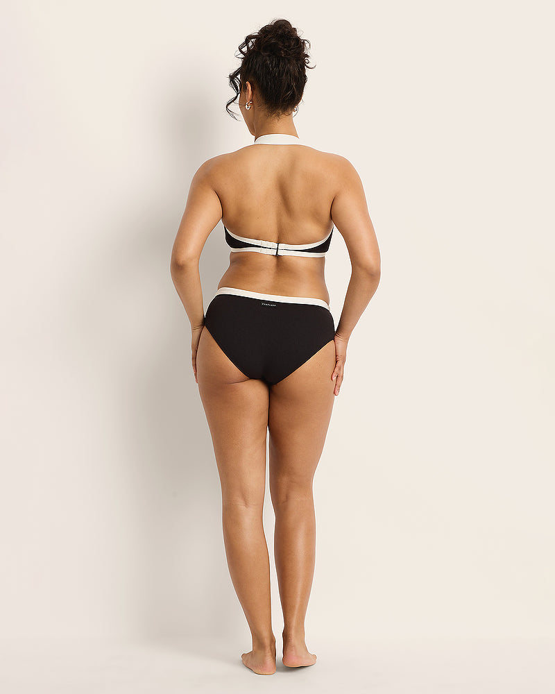 A woman stands barefoot facing away, wearing a black and white bikini. Her hair is up in a curly bun, and she is positioned against a plain, light-colored background.